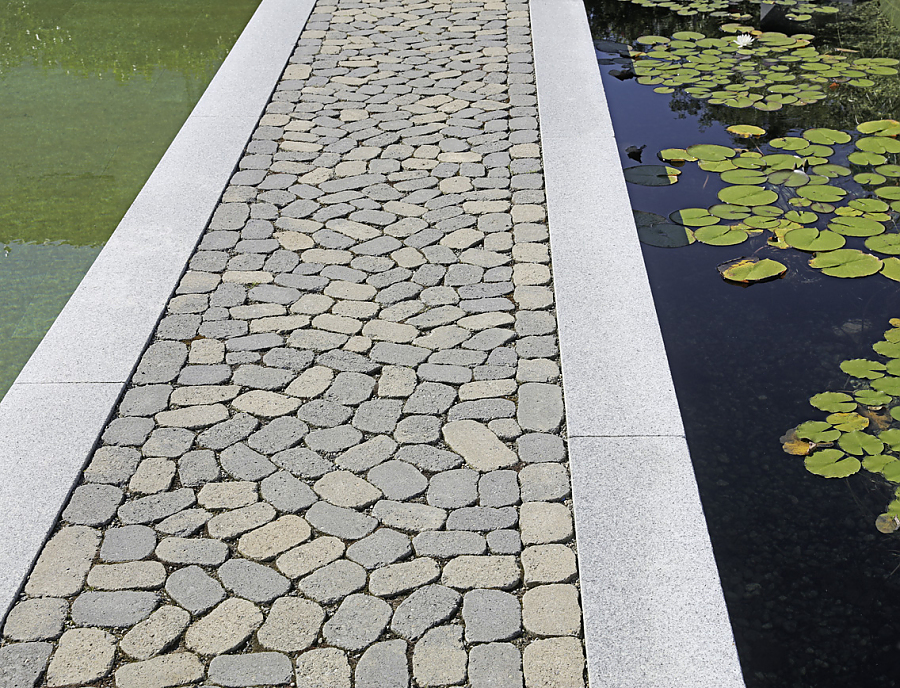 ARENA Pflastersteine aus Beton in der Farbe Staufer-Schattiert als Weg zwischen einem Pool auf der linken und einem Teich auf der rechten Seite.