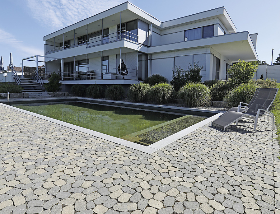 ARENA Pflastersteine aus Beton in der Farbe Melaphyr-Forte als Umrandung um einen rechteckigen Pool. Im Hintergrund ein modernes großes Haus. 