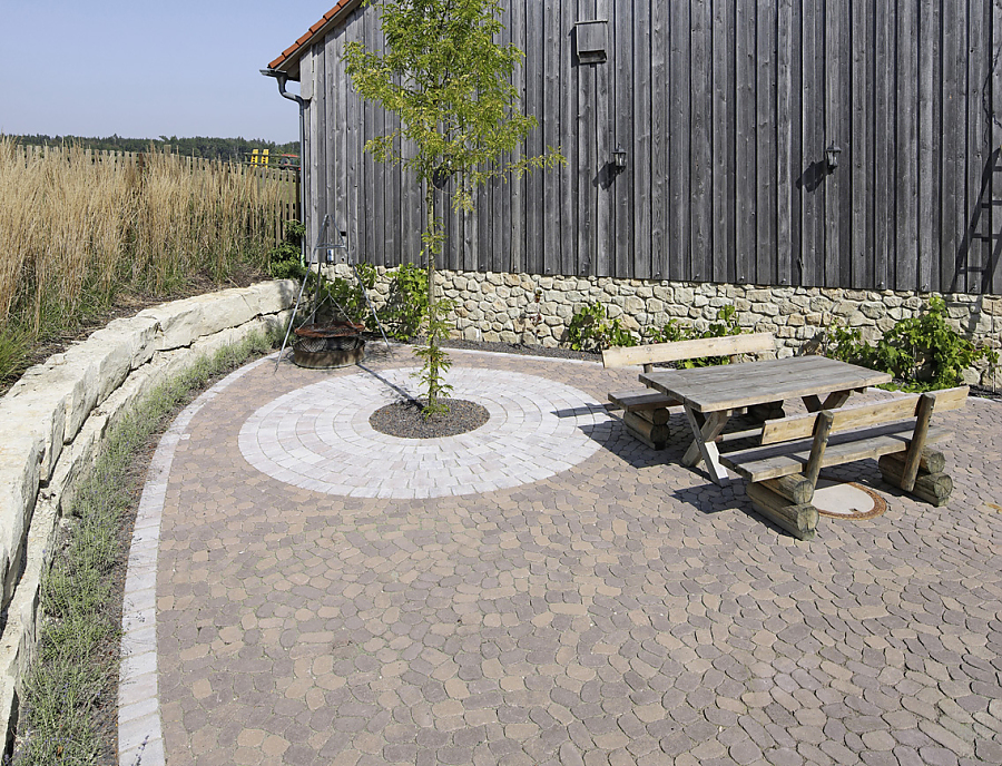 ARENA Pflastersteine aus Beton in einem Hof in der Farbe Braun-Schattiert. Darauf stehen zwei Holzbänke und einen Holztisch. Im Hintergrund ist ein Holzhaus.