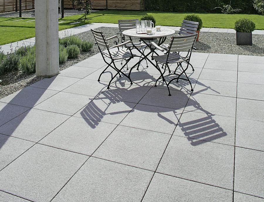ARCADO Terrassenplatten aus Beton verlegt auf einer Terrasse. Auf der Terrasse sind Stühle und ein Tisch aufgestellt, im Hintergrund sieht man den Garten.