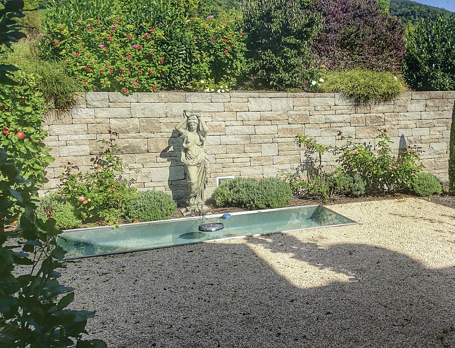 Gartenmauer SANTURO Landhausmauer in der Farbe Kalkstein-Schattiert im Garten. Im Vordergrund eine farblich passende Statue und ein Teich.