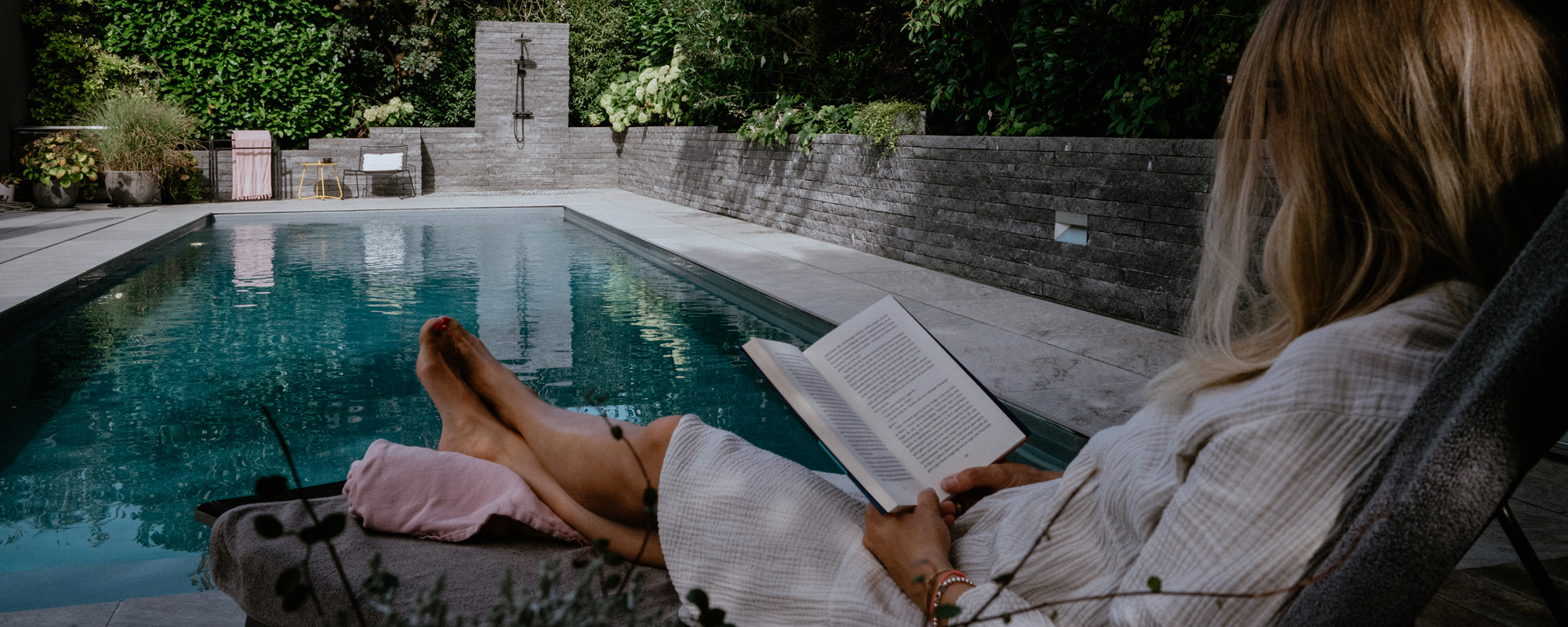 Eine Frau sitzt am Pool auf einer Liege und liest ein Buch.